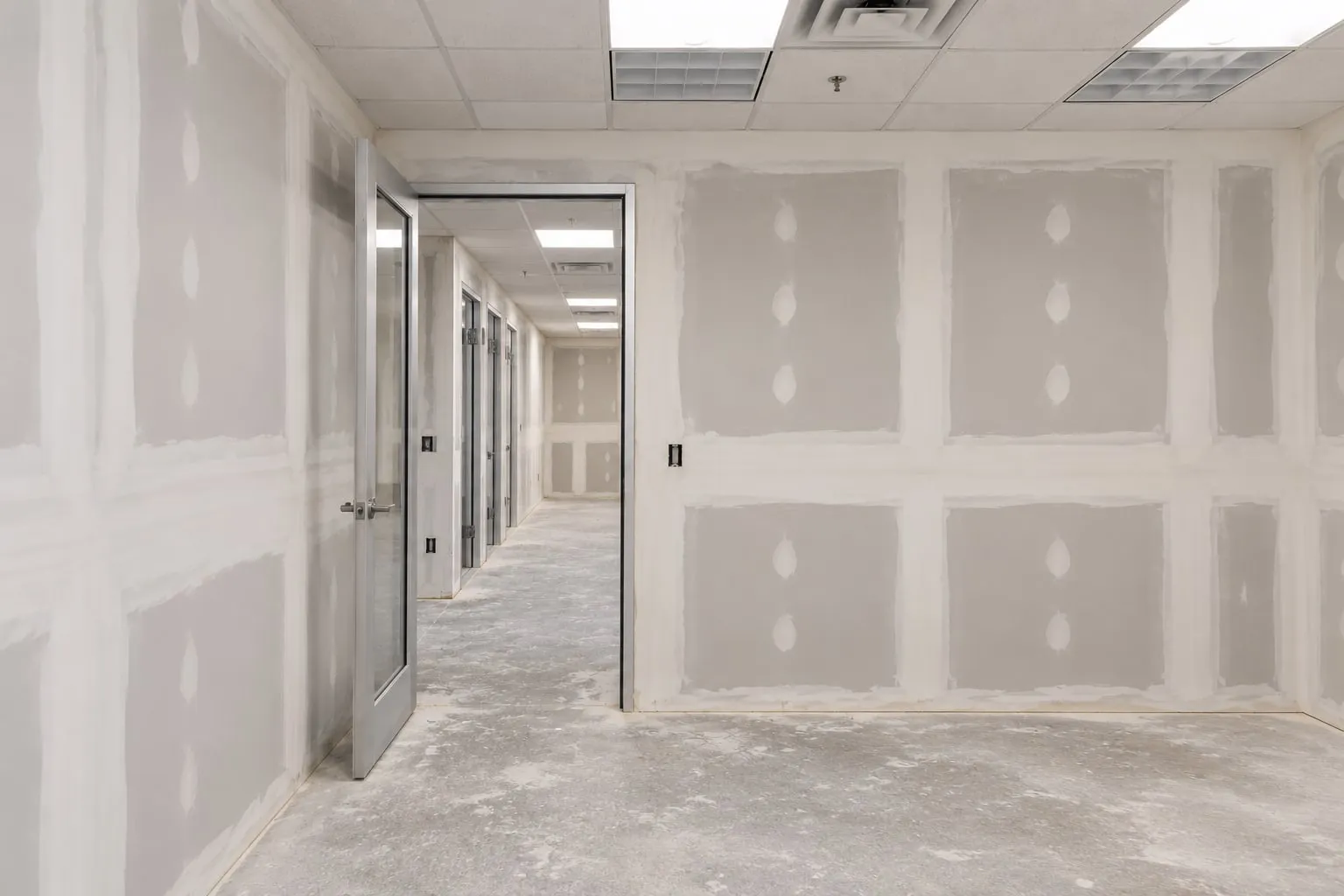 Office build-out mid-job: a private office and corridor hung and first-coated, glazed door open, acoustic ceiling in